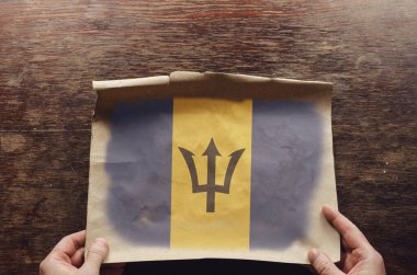 Barbados 'un sarı ve mavi bayrağı eski kağıt üzerinde. Bir adamın elleri parşömen kağıdını kahverengi bir tahta masaya bastırıyor. Yukarıdan görüntüle