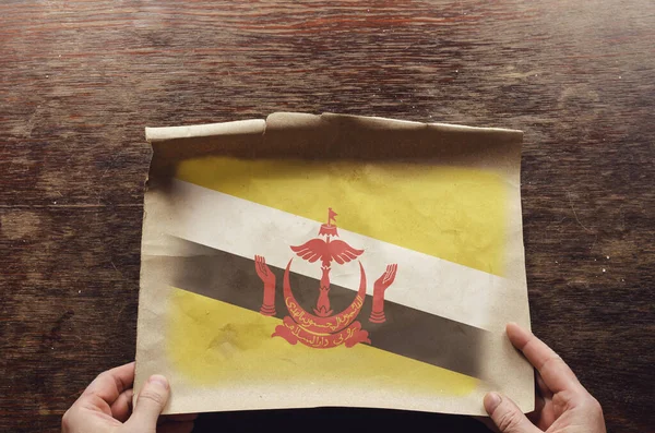 ブルネイの国旗が破れた紙の上に描かれている 男の手は茶色の木製のテーブルに羊皮紙の展開シートを押す 上からの眺め — ストック写真