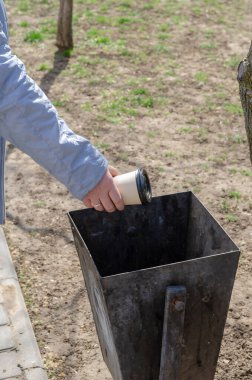 Mavi ceketli bir kadın kaldırımın yanındaki çöp kutusuna tek kullanımlık bardak atıyor. Parmağında alyansı olan yetişkin bir kadının eli, içinde kapağı olan bir kağıt bardağı vazoya koyar..