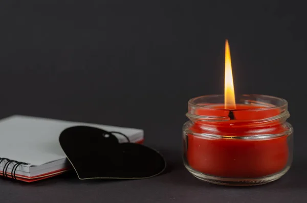 红色的蜡烛 记事本和灰色背景上的黑色心脏符号 在玻璃瓶里烧蜡蜡烛 弹跳加载的笔记本 侧视的角度 有选择的重点 — 图库照片