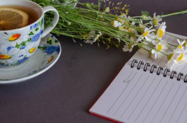 Çiçekler, bir fincan çay ve gri arka planda boş bir not defteri. Boş not defterinin üstündeki küçük papatyalar. Bir dilim limonlu seramik çay. Bir açıdan yan görünüm.