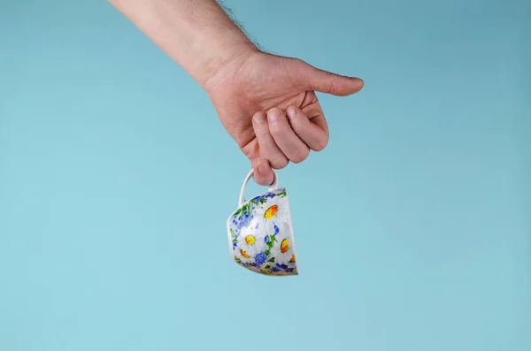 Hand and cup on blue background. Man holding White cup with painted flowers. Ceramic Mug cup for tea or coffee brightly decorated in blue and yellow colours. White daisies on the dishes.