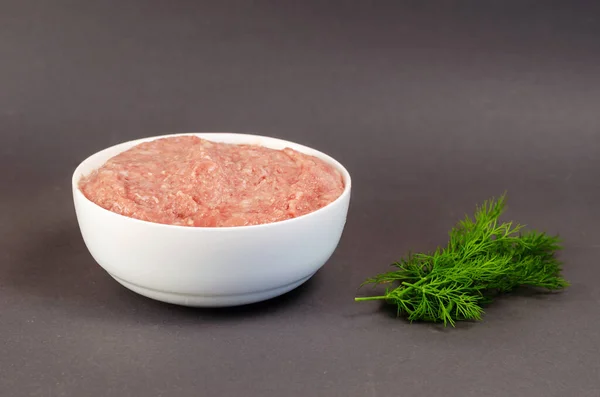 Raw ground meat and green dill on a gray background. White ceramic bowl with minced turkey. Food, ingredients. Side view.