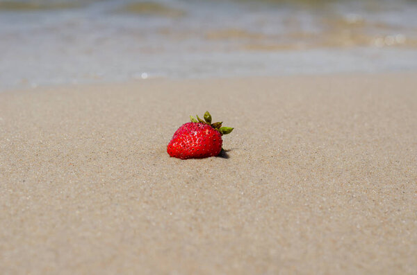 A ripe red strawberry on the wet yellow sand. The strawberry lies on the beach, washed by the wave. Selective focus.