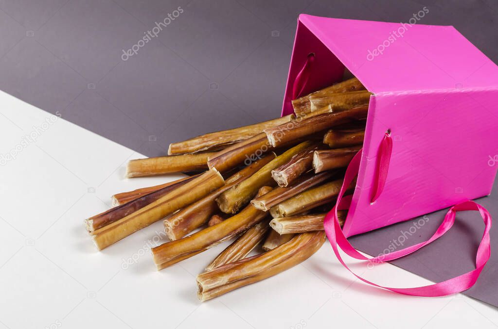 Beautiful pink gift box with pet treats. Bully sticks for dogs on gray and white backgrounds spilled out of the box lying on its side. Popular chewing treats. Beef pizzle.