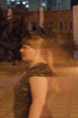 Koyu renk elbiseli, uzun saçlı yetişkin kadın portresi. Şehir binalarının önünde yürüyen saydam kadın çizgileri. Gece hayatı. Hareket bulanıklığı.