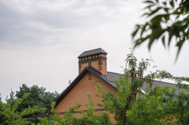 Yeşil ağaçların arasındaki kulübe. Penceresi, olukları, gelgitleri ve bacası bulutlara karşı olan kahverengi metal kiremitlerden yapılmış tuğladan bir ev. Modern yapı, mimari.