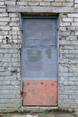 The old front door in the brick wall. A cracked front of a building with a gray-red nailed wooden door. Abandoned buildings, ruins.