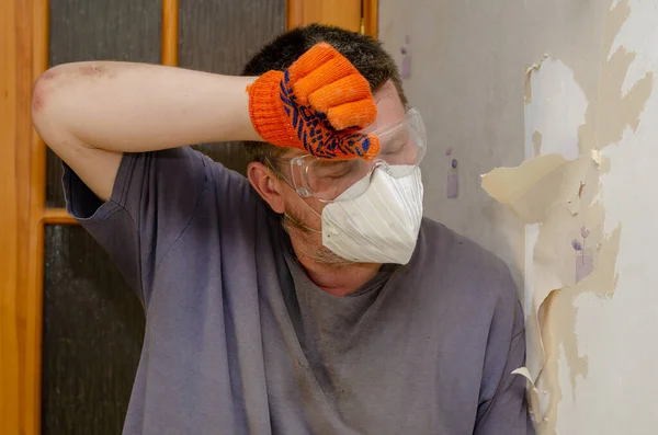 An adult male is wiping sweat from his forehead leaning against the wall. Tired male in respirator, goggles, orange gloves. Repair, Removal of paper wallpaper. Inside the room. Series part.