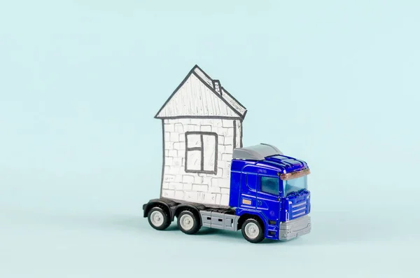 Toy tractor-trailer carrying a house. Blue plastic truck and house drawing on white paper. Blue background. Real estate, industry, transportation. Selective focus.