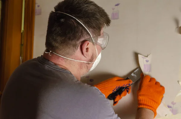 An adult man removes old wallpaper from the wall. The male is wearing a respirator, safety eyeglasses, and orange gloves. Dismantling paper wallpaper. Inside the room. Series part. Selective focus.