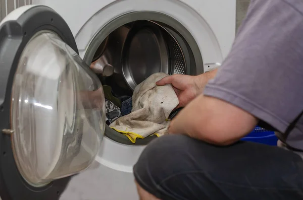 A grown man puts dirty laundry in the washing machine. A hand ho