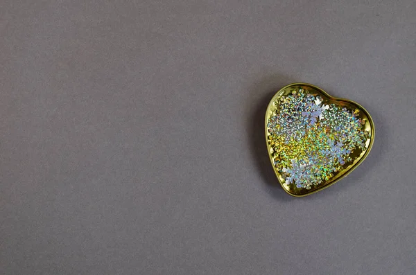 A gold metal heart filled with multicolored sequins on a gray ba