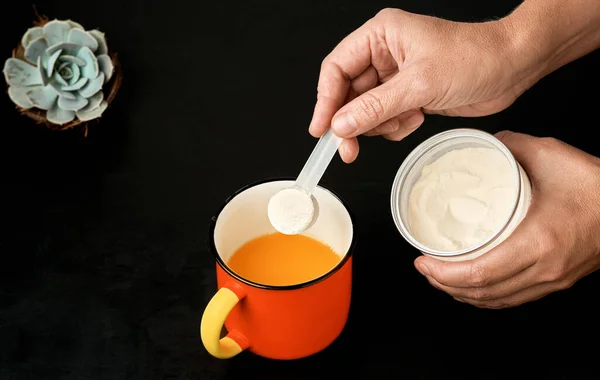 Collagen protein powder from a plastic measuring spoon female hands pour into a mug with juice, dark background, top view. Supplemental protein supplement on a natural basis for body and joint health.