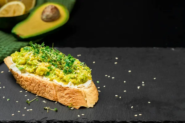 Delicious avocado toast with olive oil, tomatoes and microgreens and sesame seeds on baguette toast with cream cheese. Close-up with copy space, half an avocado next on black shale background