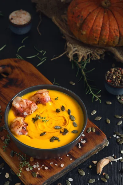 黒い木製のテーブルの上に黒いボウルにクリームとカボチャの種とソテーベーコンスライスとクリーミーなカボチャのスープ トップビュー 選択的フォーカス テーブルの上に季節のカボチャのスープを作るための成分 — ストック写真