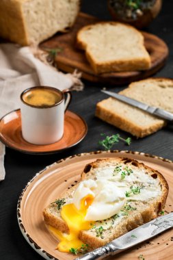 Yağlı krema ve otlarla kaplı ekmek diliminde kaynamış yumurta, siyah ahşap masadaki kil tabakta. Espresso kahve ve arka planda dilimlenmiş ekmek. Kahvaltı fikri.