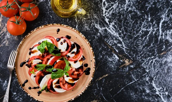 Tomatoes and mazzarella, Italian Caprese salad with tomatoes, mozzarella cheese, basil balsamic vinegar and olive oil. Dark marble background, top view, Vegetarian dish