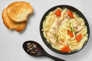 Ev yapımı tavuklu şehriye çorbası, sebze ve baharatlarla dolu bir kâse, üst manzara, beyaz beton arka plan. Sağlıklı gıda konsepti. Masadaki tahta kaşıkta ekmek ve baharatlar.