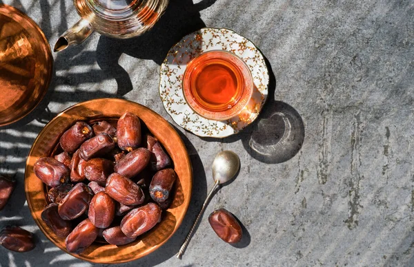 Dates on copper plate, next to red tea in glass of armudu and teapot. Layout of sweet date fruits on stone background, bright sunlight, shadows from palm leaves on table, top view with copy space
