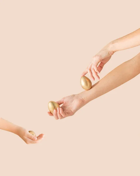Young woman\'s and children\'s hands with gold Easter eggs on the pastel beige background with copy space. Minimal creative, festive, luxury concept.