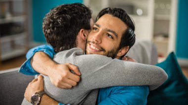 Sevimli Çekici Erkek Eşcinsel Çift Evdeki Kanepede Birlikte Oturur. Erkek arkadaşlar birbirlerine sarılıp kucaklaşıyorlar. Mutlu ve gülümsüyorlar. Gelişigüzel giyinmişler ve modern bir iç mekanı var..