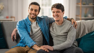 Sevimli Çekici Erkek Eşcinsel Çift Evdeki Kanepede Birlikte Oturur. Erkek arkadaşı partnerlerine el kaldırıyor. Mutlu ve gülümsüyorlar. Gelişigüzel giyinmişler ve odalarının modern bir iç mekanı var..