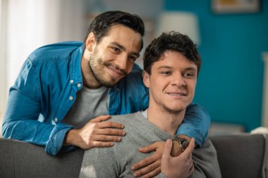 Evde Sevimli Eşcinsel Bir Çiftin Portresi. Genç adam koltuğa oturur, ortağı arkadan gelir ve onu nazikçe kucaklar. Mutlu ve gülümsüyorlar. Odanın Modern İçi Var.