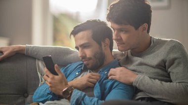 Sevimli erkek eşcinsel çift evde vakit geçiriyor. Bir kanepeye uzanıyorlar ve akıllı bir telefon kullanıyorlar. İnternette Ciddi Yüzler 'e bakıyorlar. Partners Hand onun sevgilisinin yanında. Odanın Modern İçi Var.