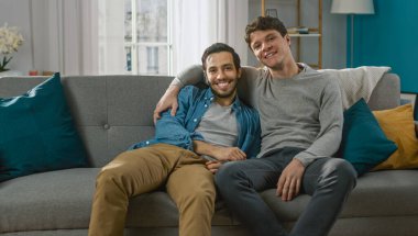 Sevimli Çekici Erkek Eşcinsel Çift Evdeki Kanepede Birlikte Oturur. Erkek arkadaşlar birbirlerine sarılıp kucaklaşıyorlar. Mutlu ve gülümsüyorlar. Gelişigüzel giyinmişler ve modern bir iç mekanı var..