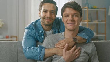 Evde Nazik Eşcinsel Bir Çiftin Portresi. Genç adam bir koltuğa oturur, ortağı onu arkadan kucaklar. Mutlu ve gülümsüyorlar. Odanın Modern İçi Var.