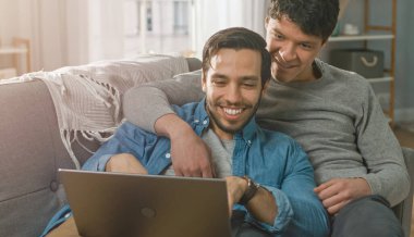 Sevimli erkek eşcinsel çift evde vakit geçiriyor. Bir kanepeye uzanıp dizüstü bilgisayarı kullanıyorlar. İnternete göz atıyorlar. Partners Hand onun sevgilisinin yanında. Gülümsüyorlar ve gülüyorlar. Odanın Modern İçi Var.
