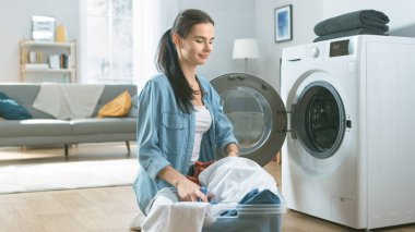 Gülümseyen Güzel Esmer Kadın, Çamaşır Makinesinin Önünde Sade Kotlarla Oturuyor. Çamaşır makinesini Kirli Çamaşırlarla dolduruyor. Modern İçişleri ile Parlak ve Geniş Oturma Odası.