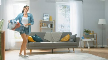 Güzel ve mutlu esmer bir kadın. Çamaşır makinesine doğru gidiyor. Üzerinde de güzel kotlar var. Çamaşır sepetini çamaşır makinesine taşıyor. Modern İçişleri ile Parlak ve Geniş Oturma Odası.