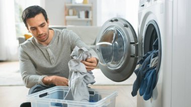 Gri Kot Giyen Yakışıklı Genç Adam ve Evde Çamaşır Makinesinin Önünde Jumper Oturuyor. Çamaşır makinesini Kirli Çamaşırlarla dolduruyor. Modern İçişleri ile Parlak ve Geniş Oturma Odası.