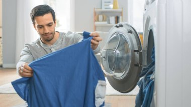 Gri Kot Giyen Yakışıklı Genç Adam ve Gömlek Evde Çamaşır Makinesinin Önünde Oturuyor. Çamaşır makinesini Kirli Çamaşırlarla dolduruyor. Modern İçişleri ile Parlak ve Geniş Oturma Odası.