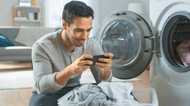 Gri Kot ve Paltolu Yakışıklı Genç Adam Çamaşır Makinesinin Önünde Oturuyor ve Akıllı Telefonunda Oyun Oynuyor. Çamaşır makinesini Kirli Çamaşırlarla dolduruyor. Modern İçişleri ile Parlak Oturma Odası.