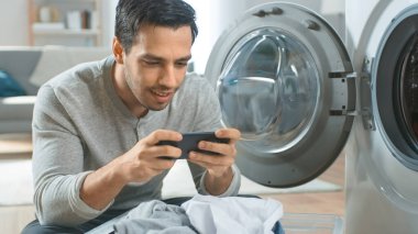 Gri Kot ve Paltolu Yakışıklı Genç Adam Çamaşır Makinesinin Önünde Oturuyor ve Akıllı Telefonunda Oyun Oynuyor. Çamaşır makinesini Kirli Çamaşırlarla dolduruyor. Modern İçişleri ile Parlak Oturma Odası.