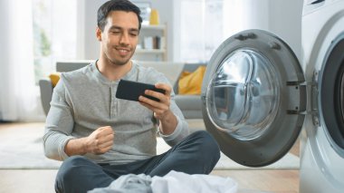 Gri Kot Giyen Yakışıklı Genç Adam Çamaşır Makinesinin Önünde Oturuyor, Akıllı Telefonuyla Oyun Oynuyor. Kazandı. Çamaşır makinesini Kirli Çamaşırlarla dolduruyordu. Modern İçişleriyle Oturma Odası.