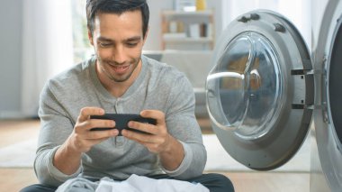 Gri Kot ve Paltolu Yakışıklı Genç Adam Çamaşır Makinesinin Önünde Oturuyor ve Akıllı Telefonunda Oyun Oynuyor. Çamaşır makinesini Kirli Çamaşırlarla dolduruyor. Modern İçişleri ile Parlak Oturma Odası.