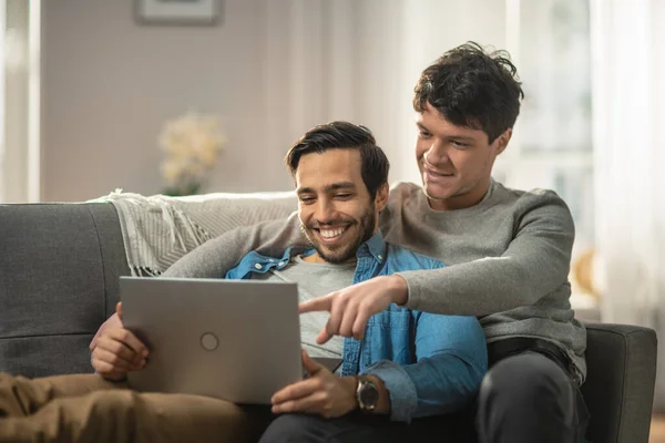 Sweet Male Queer Couple Spend Time at Home. They are Lying Down on Sofa and Use Laptop. They Browse Online. Partners Hand is Around His Lover with Other Pointing on Screen. Room Has Modern Interior.