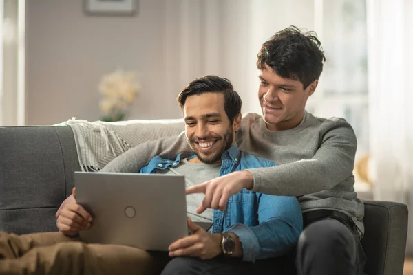 Édes férfi meleg pár otthon tölti az idejét. Lefekszenek a kanapéra és laptopot használnak. Böngészik az interneten. Partnerek kéz körül szeretője más mutatva képernyőn. A szoba modern belsővel rendelkezik. — Stock Fotó