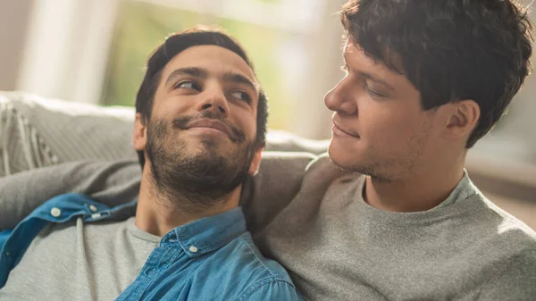 Sweet Male Queer Couple tilbringer tid hjemme. De ligger på en sofa og ser på hverandre. Hans hånd er rundt hans elsker. Det er glitrende. Rommet har moderne indre.. – stockfoto