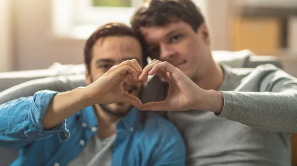 Söpön homoparin muotokuva kotona. He istuvat sohvalla ja tekevät sydänsymbolin käsillään. Kumppanilla on toinen käsi rakastajansa takana. Huone on moderni sisustus. — kuvapankkivalokuva