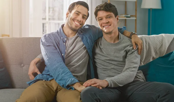 Söpö houkutteleva mies homo pari istua yhdessä sohvalla kotona. Poikaystävä laittaa kätensä kumppaneille ja he halaavat. He ovat iloisia ja hymyileviä. Ne ovat rennosti pukeutunut ja huone on moderni sisustus. — kuvapankkivalokuva