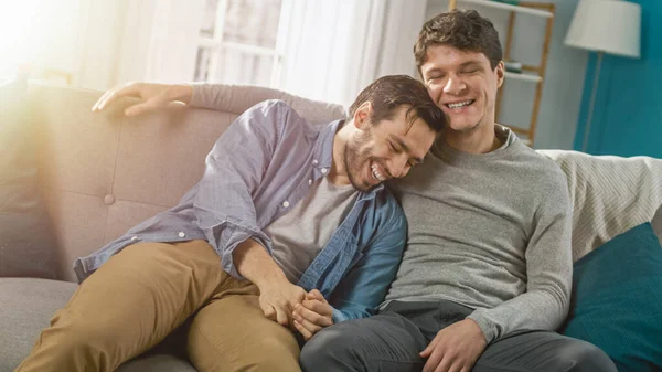 Bonito atraente masculino gay casal sentar juntos em um sofá em casa. Os namorados estão abraçando e abraçando uns aos outros. Eles estão alegres e rindo. Eles são Casualmente Vestidos e Quarto Tem Interior Moderno. — Fotografia de Stock