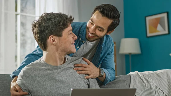 Sweet Male Gay Couple tilbringer tid hjemme. Den unge mannen arbeider på en Laptop, hans partner kommer bakfra og omfavner ham forsiktig. De ler. Rommet har moderne indre.. – stockfoto