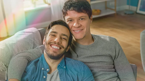 Portrait of Cute Male Queer Couple at Home. They Sit on Sofa and Look at Camera. Partner Embraces His Lover from Behind. They are Happy and Smiling. Room Has Modern Interior with Rainbow Flare Effect — Stock Photo, Image