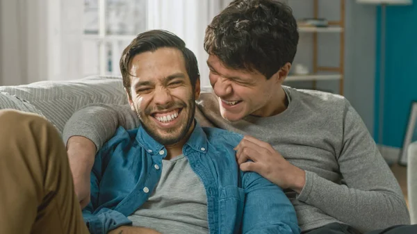 Söpön homoparin muotokuva kotona. He istuvat sohvalla ja vitsailevat. Kumppani syleilee rakastajaansa takaapäin. He ovat iloisia ja hymyileviä. Huone on moderni sisustus. — kuvapankkivalokuva