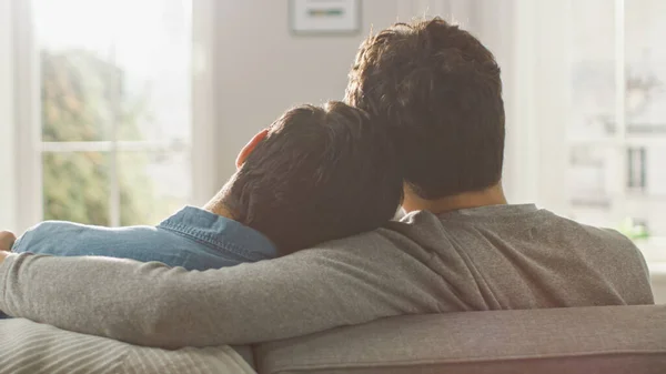 Задняя часть симпатичной мужской гей-пары обнимается, сидя на диване и складывая головы вместе. Они случайно одеты в джинсы и свитер. Номер с современным интерьером и светлым и солнечным. — стоковое фото